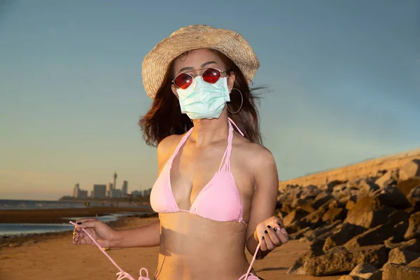 A portrait senior Asian woman wearing a pink bathing suit, wearing glasses and wearing a straw hat, with a nose mask, anti-virus COVID-19, and anti-PM2.5 dust posing happily  on the beach in evening on a blue sky.