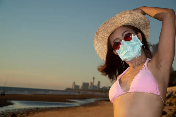 A portrait senior Asian woman wearing a pink bathing suit, wearing glasses and wearing a straw hat, with a nose mask, anti-virus COVID-19, and anti-PM2.5 dust posing happily  on the beach in evening on a blue sky.