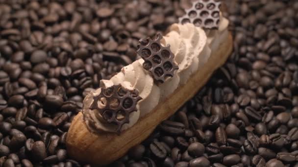 Eclair met koffiemelk en stukjes chocolade ligt op de koffiebonen — Stockvideo