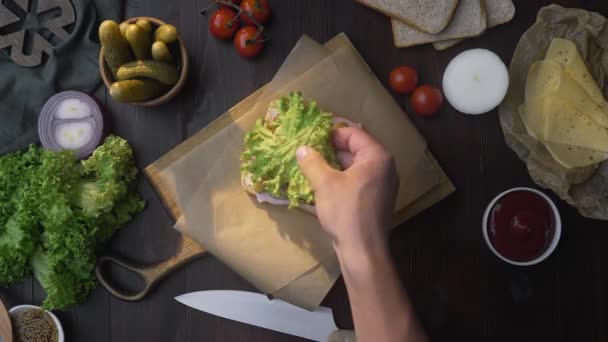 Hacer el sándwich con tocino en rodajas y ensalada chef añade ensalada al sándwich en la tabla de madera — Vídeo de stock