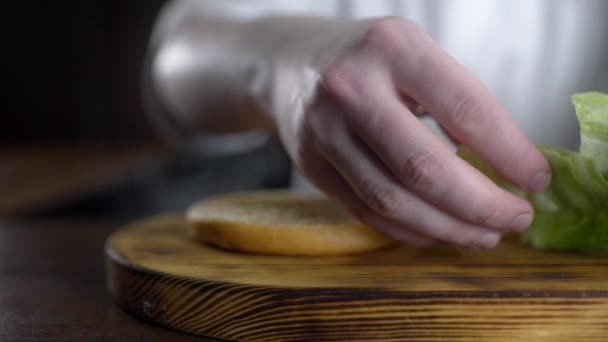 Chef cocina hamburguesa y pone hojas de ensalada verde fresco al bollo — Vídeos de Stock