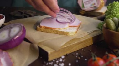 Aşçı sandviçe dilimlenmiş kırmızı soğan ve ahşap tahtaya da sebze ve jambon ekler.