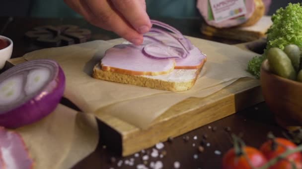 Der Koch fügt geschnittene rote Zwiebeln zum Sandwich mit Schinken und Gemüse auf dem Holzbrett hinzu — Stockvideo