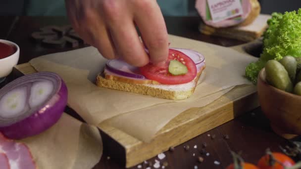 O cozinheiro adiciona picles fatiados ao sanduíche com presunto e legumes na tábua de madeira — Vídeo de Stock