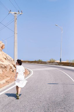 Beyaz elbiseli güzel bir kız asfalt bir yolda dağlara doğru koşuyor.