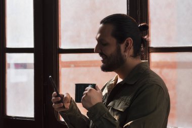 Yeşil gömlekli genç adam, pencerenin yanında telefonunu kullanırken fincandan kahve içiyor.