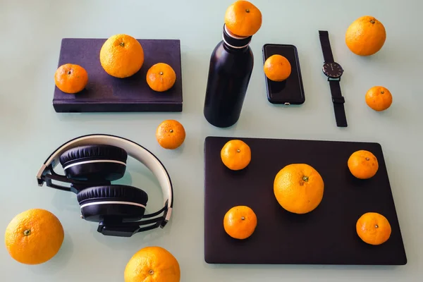 Farklı siyah nesneler, dizüstü bilgisayar, saat, şişe, telefon, kulaklık ve portakal ve mandalinalarla düzenlenmiş bir kitap..