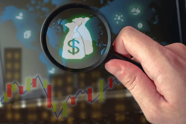Bilgisayarın önünde elinde büyüteç olan bir elin fotoğrafı. Üzerinde piyasa değeri ve para sembolleri olan bir dünya haritası var..