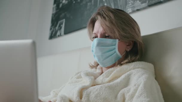 Жінка лежить на ліжку працює з ноутбуком у масці коронавірус — стокове відео