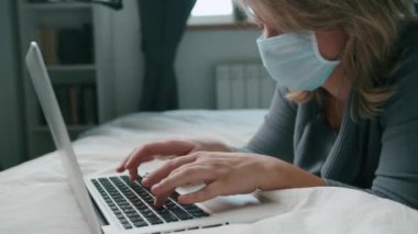 Yatakta yatan kadın, dizüstü bilgisayarla Coronavirus maskesiyle çalışıyor.