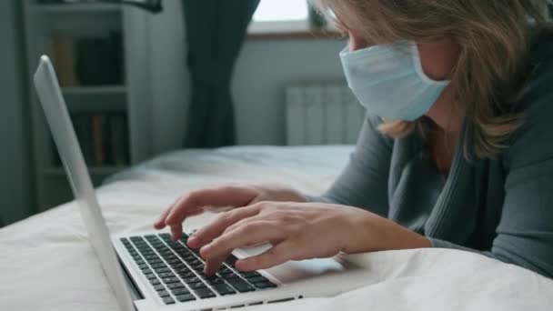 Kvinna liggande på sängen arbetar med bärbar dator i mask coronavirus — Stockvideo