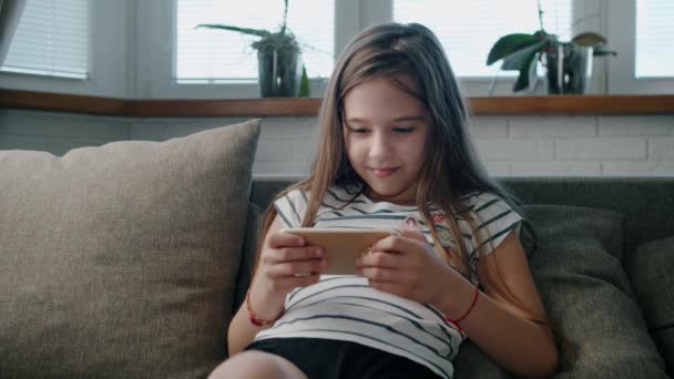 Corte hermosa niña juega en un teléfono inteligente sentado en el sofá — Vídeos de Stock