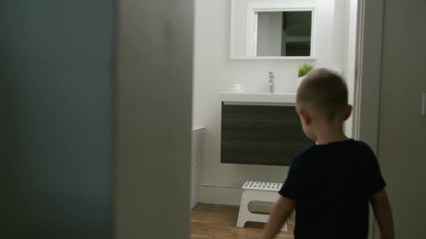 Ένα μικρό παιδί μπαίνει στο μπάνιο και ανεβαίνει σε μια καρέκλα για να πλύνει τα χέρια του. — Αρχείο Βίντεο