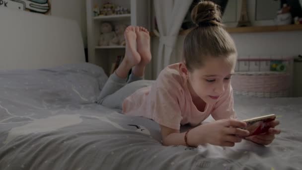 Menina bonito joga no smartphone deitado na cama — Vídeo de Stock