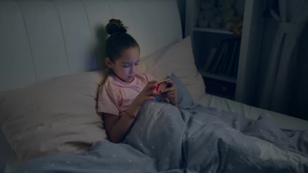 Niña linda juega en el teléfono inteligente acostado en la cama — Vídeo de stock