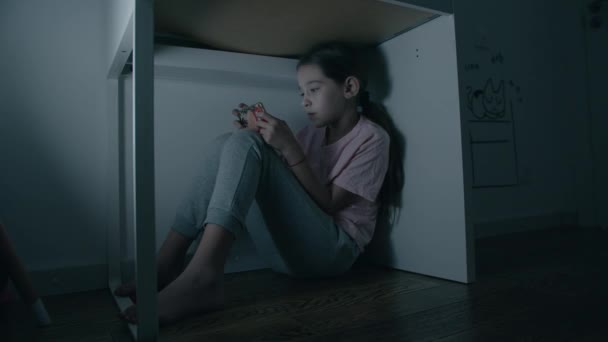 Niña emocionalmente juega en el teléfono inteligente debajo de la mesa por la noche — Vídeo de stock