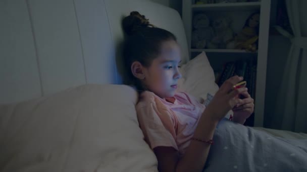 Menina bonito joga no smartphone deitado na cama — Vídeo de Stock