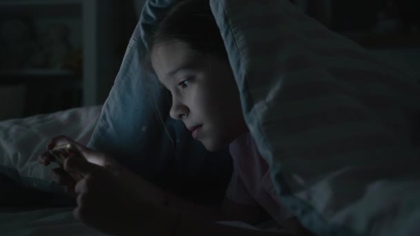 Liten flicka spelar känslomässigt i smartphone under filt på natten — Stockvideo