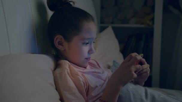 Niña linda juega en el teléfono inteligente acostado en la cama — Vídeos de Stock