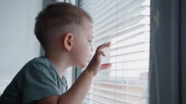 Küçük sarışın çocuk pencerenin perdelerinden dışarı bakıyor.