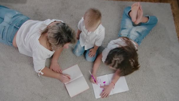 美丽的快乐的女人和年轻的女孩和男孩，妈妈，女儿和儿子画了一个笔记本躺在地板上明亮的阳光灿烂的一天。头像镜头 — 图库视频影像