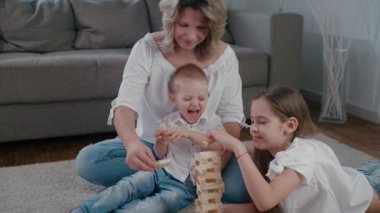 Mutlu aile annesi kız ve oğlu evde Jenga oynarken eğleniyor. Gülün, gülümseyin ve birlikte güneşli bir gün kurun. Yavaş çekim kamerası
