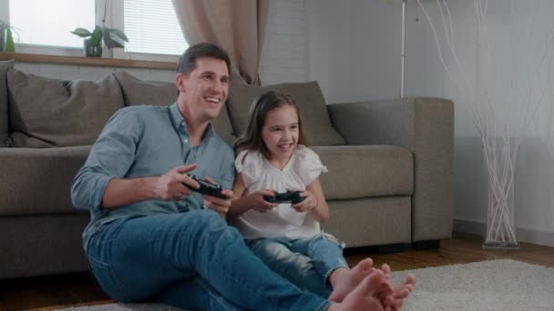 Divertido padre e hija ríen empujó y jugar un juego de computadora de la consola. La chica gana y con deleite levanta las manos. Movimiento lento — Vídeo de stock