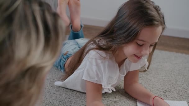 Mulher feliz bonita e jovem, mãe e filha desenhar em um caderno deitado no chão da casa dia ensolarado brilhante. Fechar e movimento lento — Vídeo de Stock