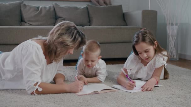Bella donna felice e giovane ragazza e ragazzo, mamma, figlia e figlio disegnare in un quaderno sdraiato sul pavimento della casa luminosa giornata di sole — Video Stock
