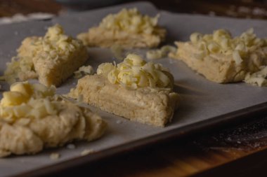 Pişirme tepsisindeki parşömen üzerinde çiğ peynirli çörekler. Çörekler rendelenmiş peynirle kaplanmış.
