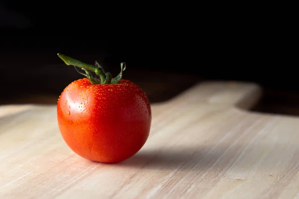 A single tomato viewed from the side sitting on a wooden board. The tomato is covered with condensation to give the appearance of freshness.