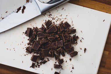 Beyaz bir doğrama tahtasının üzerinde kaba küplere bölünmüş çikolatanın yukarıdan görünüşü. Çikolatayı kesmek için kullanılan bıçak da çerçevede.