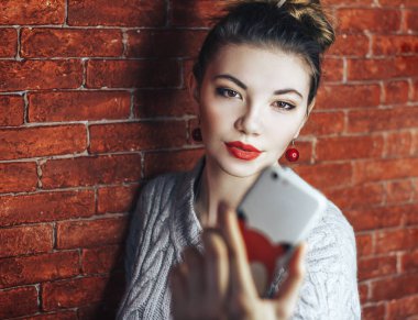 Güzel kadın duvar tuğla önünde duran ve selfie yapmak. Kırmızı dudaklar ile. Telefon odaklanmamış.