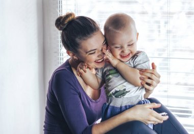 Mutlu neşeli aile. Anne ve bebek öpüşme, gülen ve sarılma. Pencere oturmak.