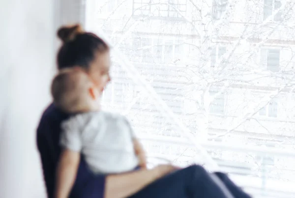 A sad mother sitting with her baby on the windowsill, looking out the window and waiting for something. The baby is lying on Mom's shoulder. Family unfocused, focus on window.