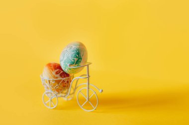 Sarı arka planda beyaz bisiklet sepetinde renkli Paskalya yumurtaları.