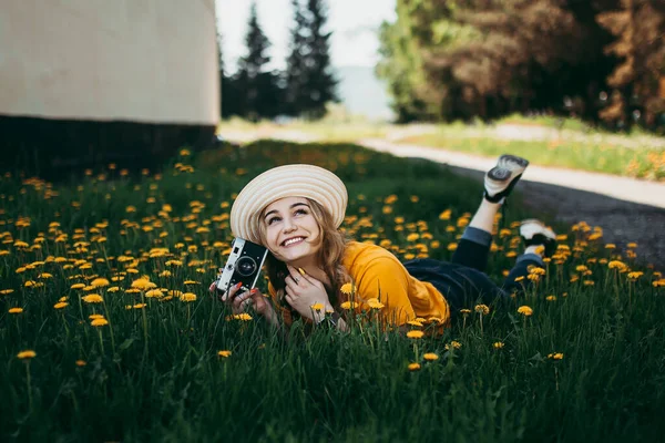 Turuncu tişörtlü genç bir kız elinde retro kamerayla çimlere uzanmış karahindiba çiçekli karahindibalar. — Stok fotoğraf