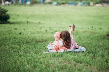 Küçük çocuklu güzel anne sıcak bir günde çimlerin üzerinde açık gökyüzünün altında pembe elbiseleriyle dinleniyor.