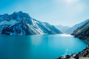 A dramatic panorama of El Yeso Dam with turquoise color and sun flares reflecting in the water in the Andes Mountains with snow on the peaks and rock parts of the mountain exposed. clipart