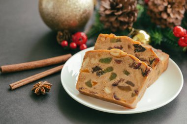 Beyaz tabakta tatlı meyveli pasta dilimleri Noel süsüsüyle yan manzaralı siyah granit masaya konur. Noel kutlaması için geleneksel romlu meyveli pasta. Ev yapımı fırın konsepti.