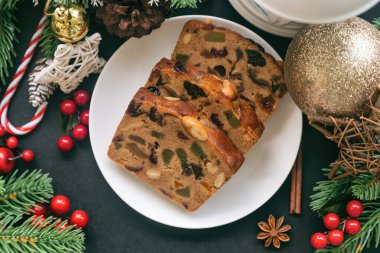 Beyaz tabakta tatlı meyveli kek dilimleri üst kattaki siyah granit masaya konulmuş ve Noel süslemesi yapılmış. Noel kutlaması için geleneksel romlu meyveli pasta. Ev yapımı fırın konsepti.