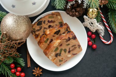 Beyaz tabakta tatlı meyveli kek dilimleri üst kattaki siyah granit masaya konulmuş ve Noel süslemesi yapılmış. Noel kutlaması için geleneksel romlu meyveli pasta. Ev yapımı fırın konsepti.