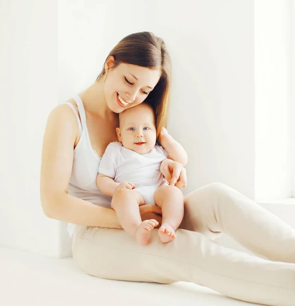 Feliz madre sonriente con bebé en casa en habitación blanca cerca de la ventana —  Fotos de Stock