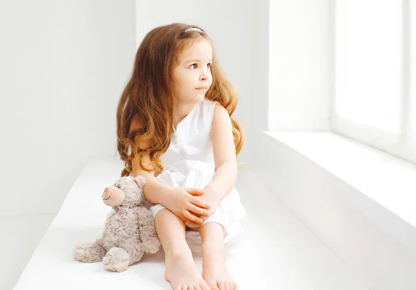 Niña pequeña con osito de peluche sentado en casa en habitación blanca —  Fotos de Stock