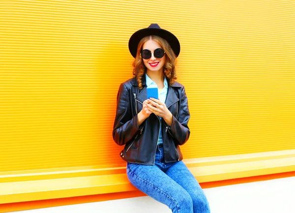 Модная улыбающаяся женщина пользуется смартфоном в черной рок-куртке — стоковое фото