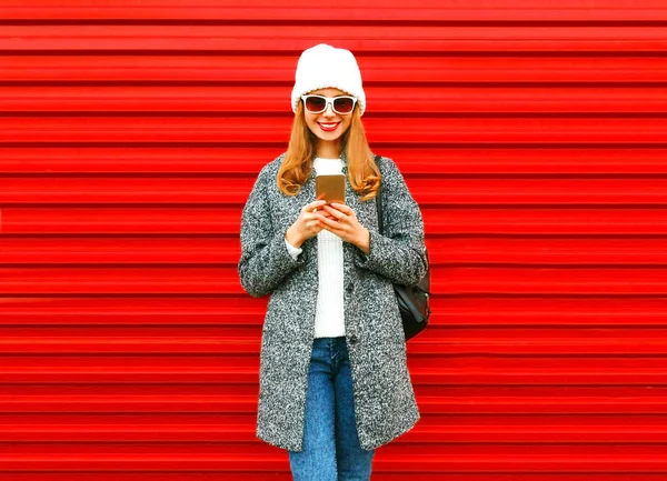 Moda sonriente mujer usando teléfono inteligente sobre un fondo rojo en th —  Fotos de Stock