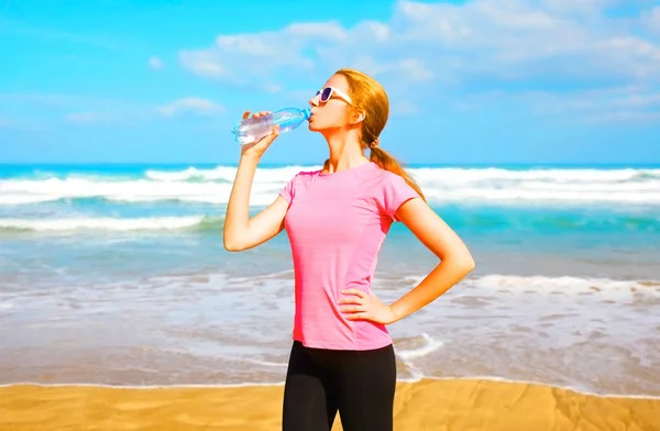 Fitness femme boit de l'eau de bouteille sur la plage près de la mer — Photo