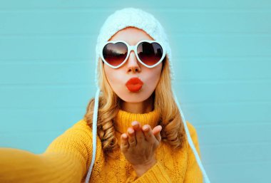 Kapalı kış portresi çek güzel kadın kırmızı dudaklar üfle tatlı hava öpücüğü ver selfie çektiği için, sarı örgü süveter, beyaz şapka, mavi arka planda kalp şeklinde güneş gözlüğü takıyor.