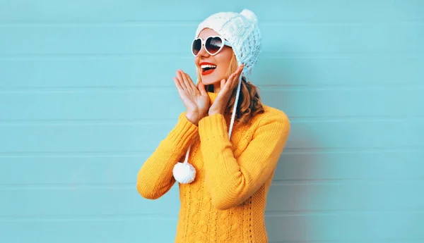 Winter portrait happy surprised young woman wearing yellow knitted sweater and white hat with pom pom, heart shaped sunglasses on blue wall background