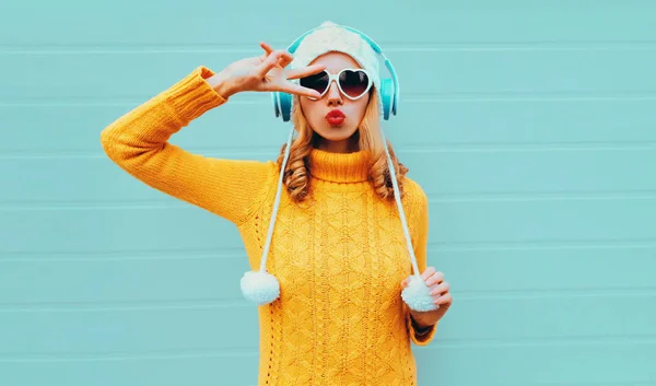 Winter portrait cool young woman in wireless headphones listening to music blowing red lips wearing yellow knitted sweater and white hat with pom pom, heart shaped sunglasses on blue wall background